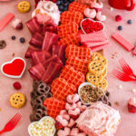 a large wooden board filled with different pink treats including pink heart shaped waffles, strawberry cinnamon rolls, and heart shaped bacon