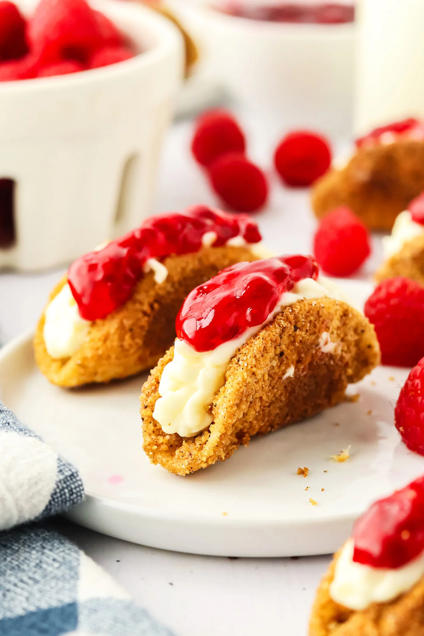 cheesecake tacos on a plate surrounded by fresh raspberries and a blue and white napkin